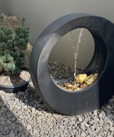 Kreisförmiger Brunnen mit Wasser, umgeben von Steinen und einer kleinen Pflanze.