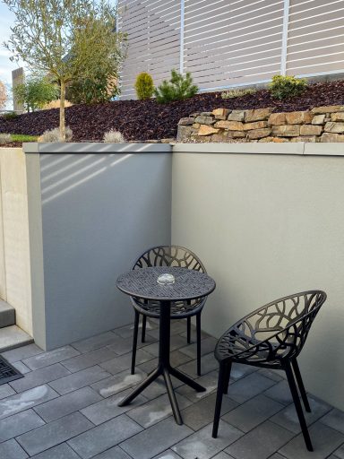 Kleiner Tisch mit zwei Stühlen in einem Außenbereich, umgeben von Wänden und Pflanzen.