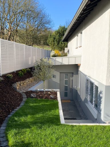 Haus mit modernem Eingangsbereich und gepflegtem Garten, umgeben von einem Zaun.